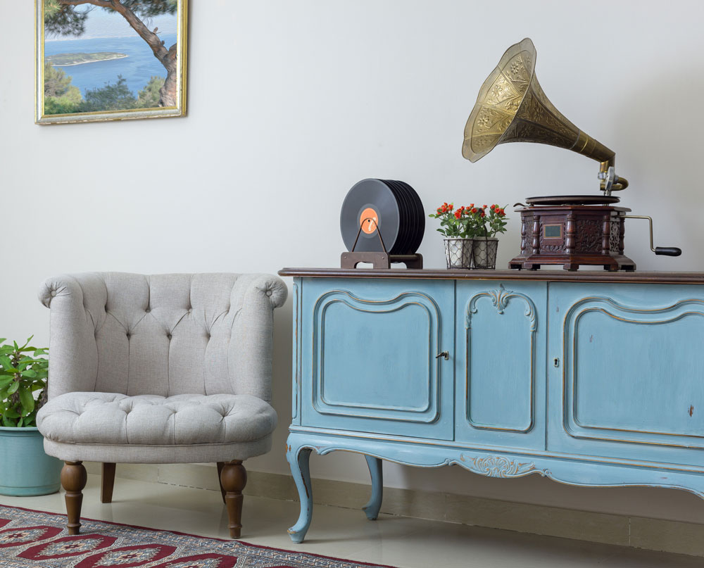 Vintage interior of retro off white armchair, vintage wooden light blue sideboard, old phonograph (gramophone) and vinyl records on background of beige wall, tiled porcelain floor, and red carpet.
