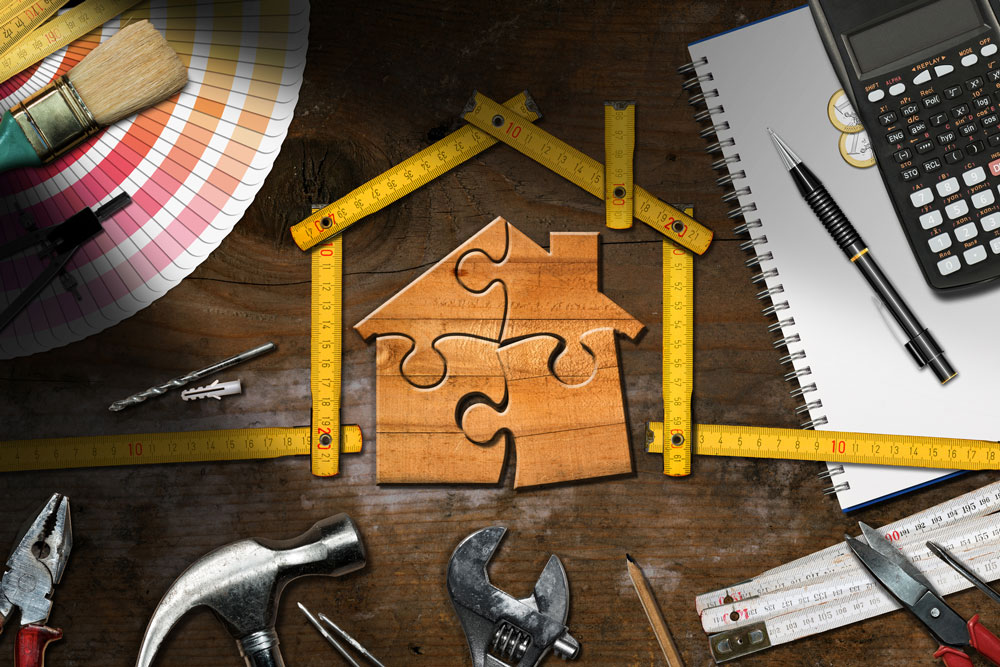 Wooden house made of puzzle pieces on a wooden workbench with work tools, folding ruler in the shape of a house, calculator and Euro coins.